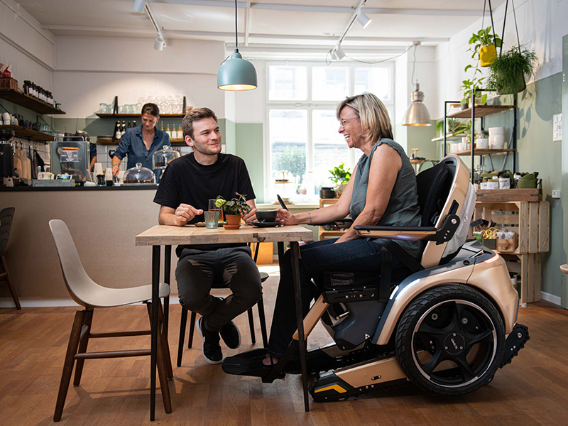 Frau sitzt mit Elektrorollstuhl Scewo Bro an Esstisch
